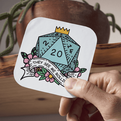 Hand holding a coaster with a D20 DnD die and text, with a plant and pot in the background