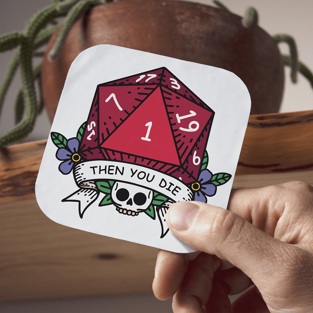 Sticker with a red DnD D20 die and skull design held by a hand, with a plant and wooden surface in the background.