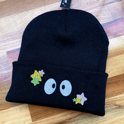 A black Soot Sprites beanie features embroidered eyes and yellow stars inspired by Ghibli's Susuwatari.