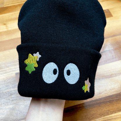 A black Soot Sprites beanie, inspired by Ghibli's Susuwatari, features large embroidered eyes and small yellow stars on a cuffed brim being worn over a hand.
