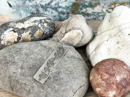 The Quirkify Star Trek Vulcan IDIC Calligraphy Sterling Silver Pendant Necklace features an intricate rectangular design, elegantly displayed on smooth stones with a softly gleaming finish against a muted background.