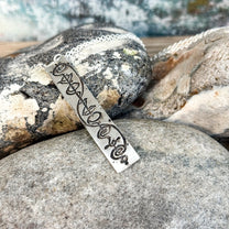The Quirkify Star Trek Vulcan IDIC Calligraphy Sterling Silver Pendant Necklace features an intricate rectangular design, elegantly displayed on smooth stones with a softly gleaming finish against a muted background.