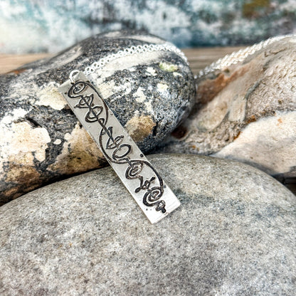 The Quirkify Star Trek Vulcan IDIC Calligraphy Sterling Silver Pendant Necklace features an intricate rectangular design, elegantly displayed on smooth stones with a softly gleaming finish against a muted background.