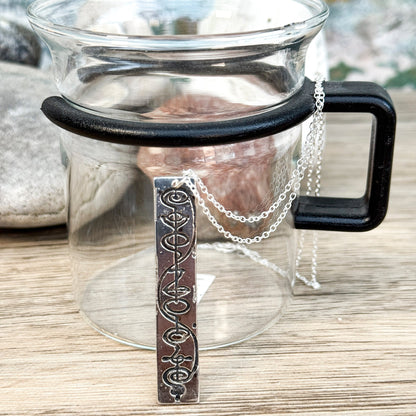 The Quirkify Star Trek Vulcan IDIC Calligraphy Sterling Silver Pendant Necklace features an intricate rectangular design, elegantly displayed on smooth stones with a softly gleaming finish against a muted background.