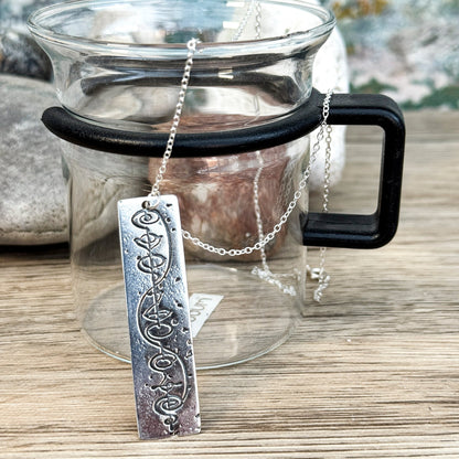 The Quirkify Star Trek Vulcan IDIC Calligraphy Sterling Silver Pendant Necklace features an intricate rectangular design, elegantly displayed on smooth stones with a softly gleaming finish against a muted background.