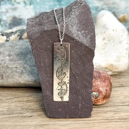 The Quirkify Star Trek Vulcan IDIC Calligraphy Sterling Silver Pendant Necklace features an intricate rectangular design, elegantly displayed on smooth stones with a softly gleaming finish against a muted background.