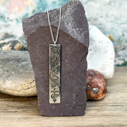 The Quirkify Star Trek Vulcan IDIC Calligraphy Sterling Silver Pendant Necklace features an intricate rectangular design, elegantly displayed on smooth stones with a softly gleaming finish against a muted background.
