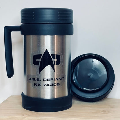 A silver Star Trek-inspired travel mug labeled "U.S.S. Defiant NX 74205" sits next to its black lid on a wooden surface.