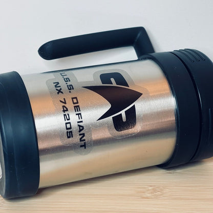 A silver Star Trek-inspired travel mug displays "U.S.S. DEFIANT NX 74205" and a white arrow graphic on its side, featuring a black handle.
