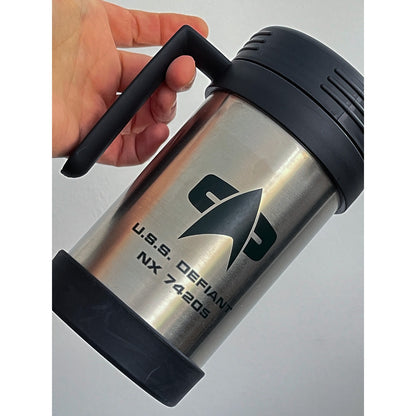 A silver Star Trek-inspired travel mug labeled "U.S.S. DEFIANT NX 74205" is held by a person's hand.