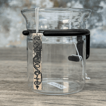 A Sterling Silver Vulcan IDIC pendant necklace hangs from a delicate chain, displayed against a glass tea infuser on a wooden surface.