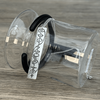 A sterling silver Vulcan IDIC calligraphy etched pendant necklace hangs from a black ribbon attached to a clear plastic display bag on a wooden surface.