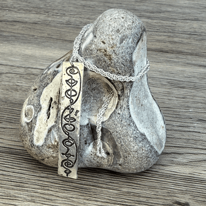 A sterling silver Vulcan IDIC calligraphy etched pendant hangs from a delicate chain against a textured stone background.