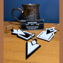 Three 3D acrylic keyrings shaped like the Skyrim quest marker icon are arranged on a wooden surface in front of a metal mug.