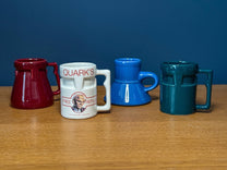 Four small ceramic mugs in burgundy, white, blue, and teal are lined up on a wooden surface, including one white mug featuring Quark's logo from the DS9 Mini Raktajino Mug Set.