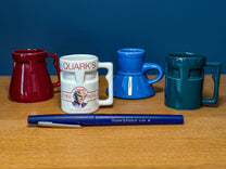Four small colorful mugs in red, white, blue, and teal sit on a wooden surface next to a blue pen, featuring graphics from the DS9 Raktajino mug set.