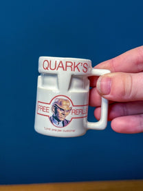 A hand holds a small white ceramic DS9 Mini Raktajino mug featuring Quark's logo and an image of a character with the text Free Refills.