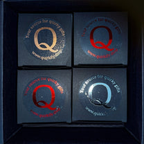 Four small black gift boxes arranged in a square, each featuring a large stylized letter Q in different metallic colors and the text Your source for quirky gifts with the website www.quirkify.shop.