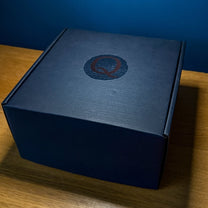 A dark blue square gift box featuring a circular emblem with a red letter Q on top.