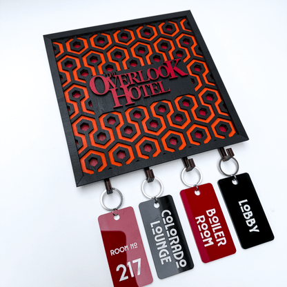 A laser-cut wooden key holder featuring "The Overlook Hotel" name and a carpet pattern design is displayed with several keychains labeled "Room 217," "Colorado Lounge," "Boiler Room," and "Lobby.