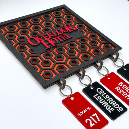 A laser-cut wooden key holder featuring a carpet pattern and "The Overlook Hotel" logo from The Shining is displayed with several red key tags labeled with room numbers and locations.