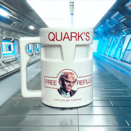 A white ceramic mug labeled "Quark's" features a portrait of Quark and text promoting free refills, set against a backdrop resembling the interior of a Star Trek: Deep Space Nine space station.