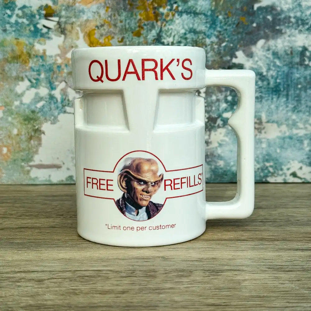 Quarks Bar Mug replica from Star Trek Deep Space Nine labelled "Quark's" with a portrait of Quark and text promoting free refills, against a colourful background