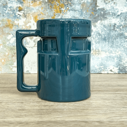 A teal Ops Raktajino mug from DS9 sits on a wooden surface against a textured blue and gold wall.