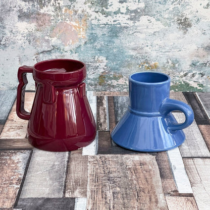 Two Raktajino Replica mugs, one red and one blue, sit on a wooden surface against a textured wall.