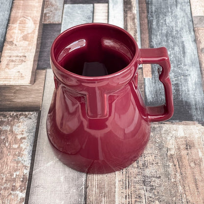 A red Raktajino replica mug resembling a Big Red Synth Ale stein from Quark’s Bar in DS9 sits on a patterned wooden surface.