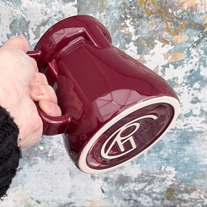 A hand holds a red Raktajino Replica Mug featuring a stylized "Q" logo on its base, referencing Quark's Bar from DS9.