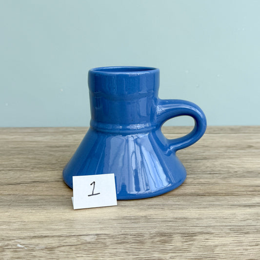A blue handmade "Rejected Raktajino Mug" by Quirkify, featuring a wide flared base and handle, sits on a wooden surface. A white paper labeled "1" is in front, with a light blue background. Give one a good home today!.