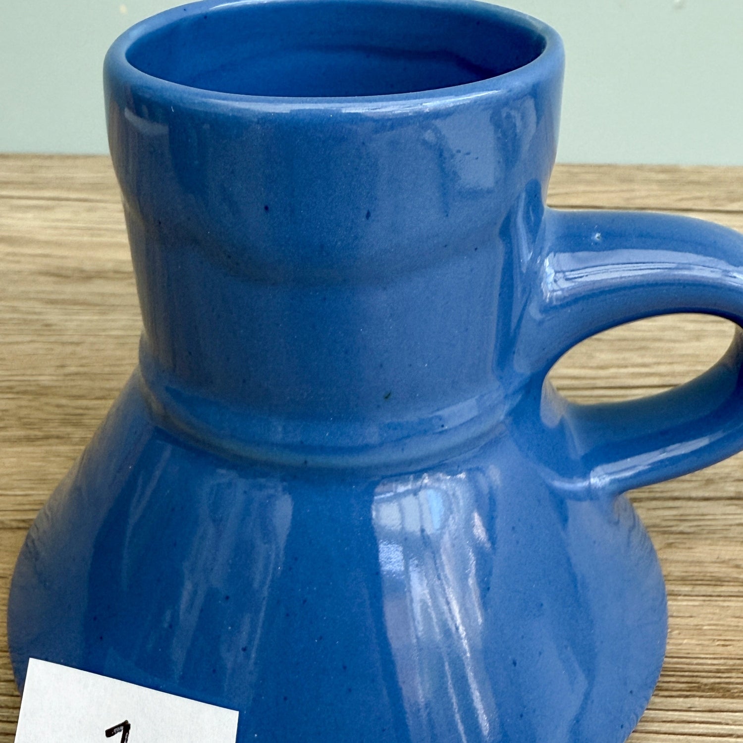 A blue handmade "Rejected Raktajino Mug" by Quirkify, featuring a wide flared base and handle, sits on a wooden surface. A white paper labeled "1" is in front, with a light blue background. Give one a good home today!.