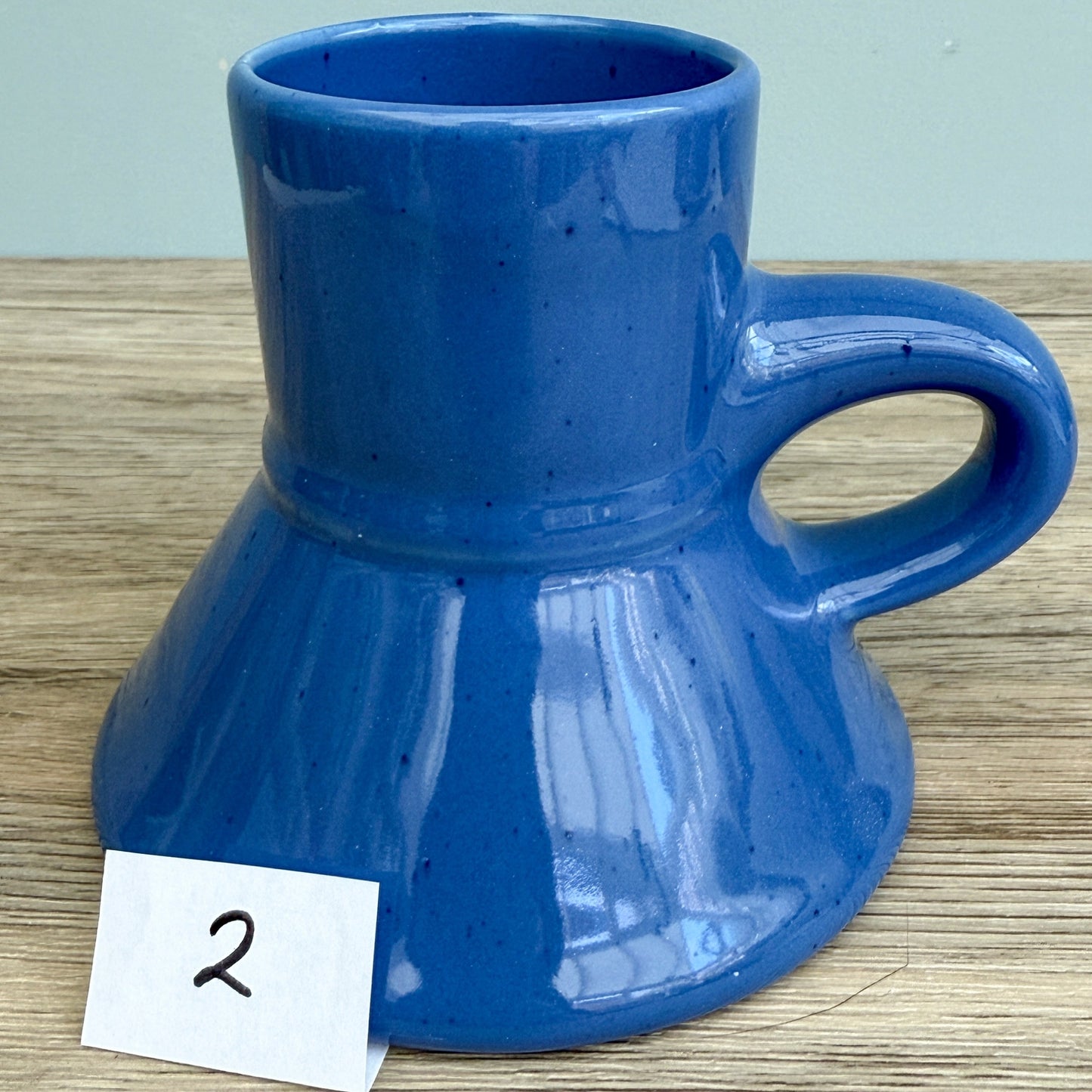 A blue handmade "Rejected Raktajino Mug" by Quirkify, featuring a wide flared base and handle, sits on a wooden surface. A white paper labeled "1" is in front, with a light blue background. Give one a good home today!.