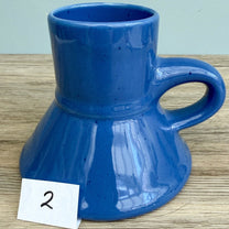 A blue handmade "Rejected Raktajino Mug" by Quirkify, featuring a wide flared base and handle, sits on a wooden surface. A white paper labeled "1" is in front, with a light blue background. Give one a good home today!.