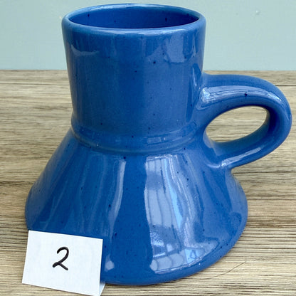 A blue handmade "Rejected Raktajino Mug" by Quirkify, featuring a wide flared base and handle, sits on a wooden surface. A white paper labeled "1" is in front, with a light blue background. Give one a good home today!.