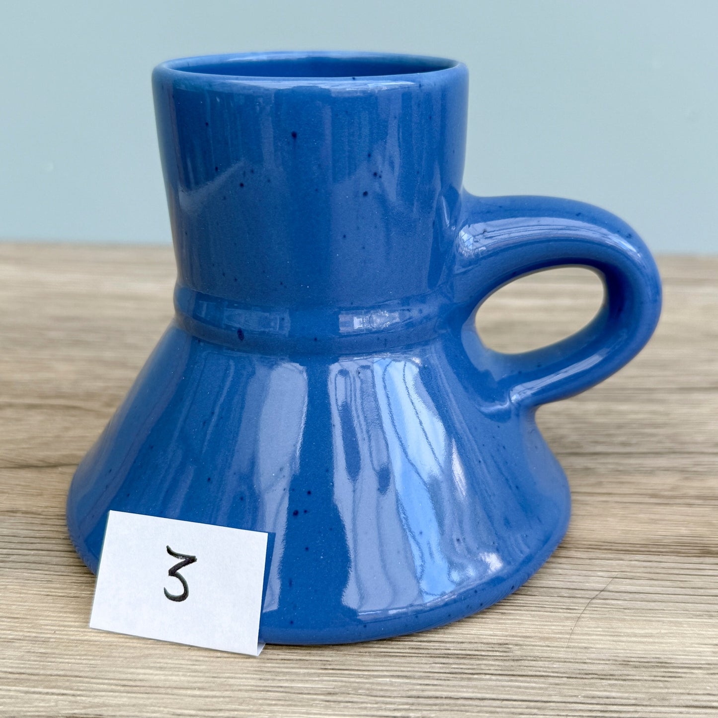 A blue handmade "Rejected Raktajino Mug" by Quirkify, featuring a wide flared base and handle, sits on a wooden surface. A white paper labeled "1" is in front, with a light blue background. Give one a good home today!.