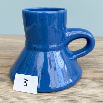 A blue handmade "Rejected Raktajino Mug" by Quirkify, featuring a wide flared base and handle, sits on a wooden surface. A white paper labeled "1" is in front, with a light blue background. Give one a good home today!.