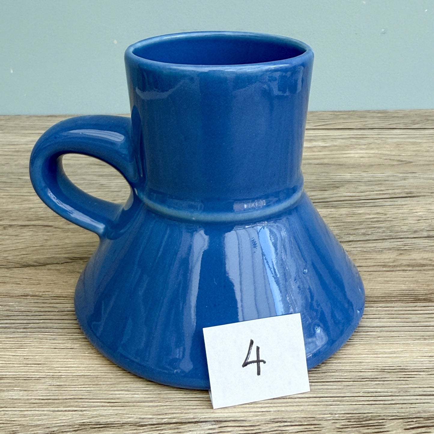 A blue handmade "Rejected Raktajino Mug" by Quirkify, featuring a wide flared base and handle, sits on a wooden surface. A white paper labeled "1" is in front, with a light blue background. Give one a good home today!.