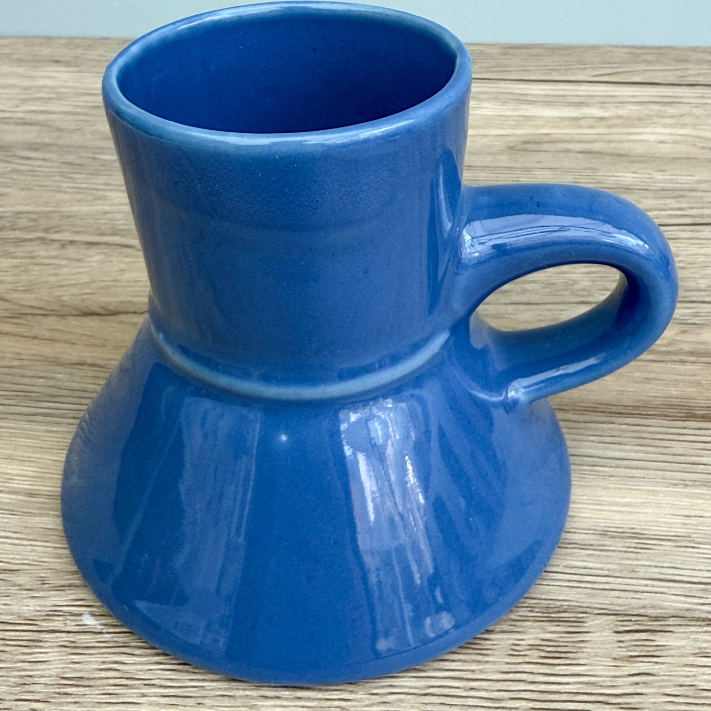 A blue handmade "Rejected Raktajino Mug" by Quirkify, featuring a wide flared base and handle, sits on a wooden surface. A white paper labeled "1" is in front, with a light blue background. Give one a good home today!.