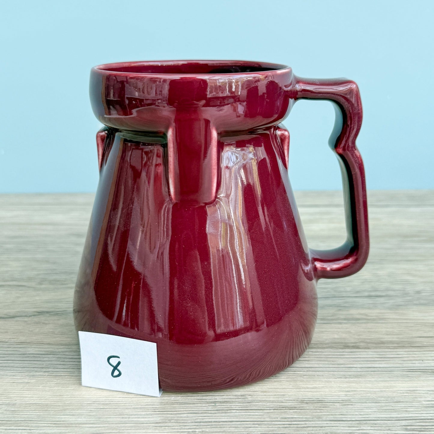 A red handmade "Rejected Raktajino Mug" by Quirkify, featuring a wide flared base and handle, sits on a wooden surface. Give one a good home today!.