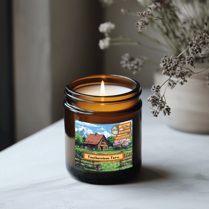 A lit Stardew Valley-themed candle in a brown jar sits on a white surface, displaying an illustration of Featherstone Farm and seasonal scent labels.