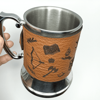 A steel beer mug is wrapped in upcycled brown leather featuring Dungeons and Dragons-inspired designs like a book, bow, and axe.