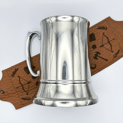 A steel beer mug is wrapped with upcycled leather featuring a Dungeons and Dragons adventurer's starter kit design of arrows, an axe, and foliage.