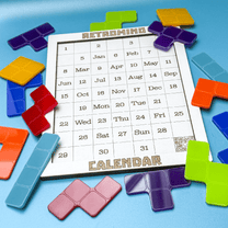 Colorful KronoBlox puzzle pieces are arranged around a calendar displaying dates and months, referencing Tetromino shapes for a perpetual calendar design.