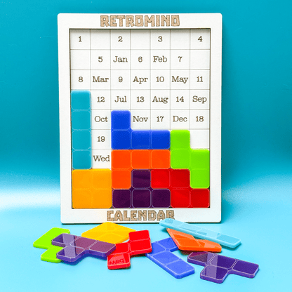 A white KronoBlox puzzle calendar displays dates and months arranged as Tetromino shapes alongside loose colorful blocks on a blue surface.