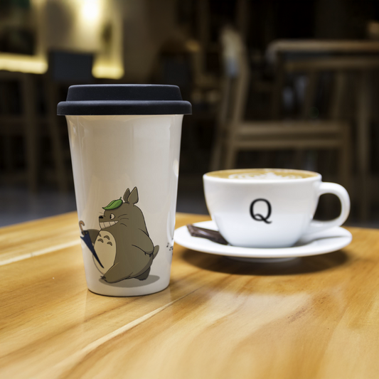 Ceramic travel mug with Totoro from Studio Ghibli and a Quirkify coffee cup on a wooden table.