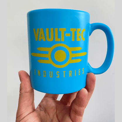 A blue Fallout mug displays the Vault-Tec Industries logo in yellow lettering and design.