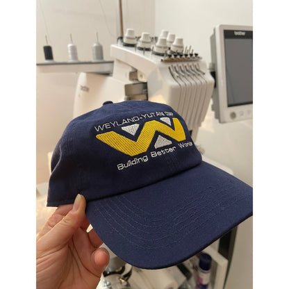 A navy blue baseball cap features an embroidered Weyland-Yutani Corp logo with a stylized "W" and the phrase "Building Better Worlds.
