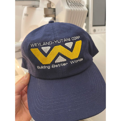 A navy blue baseball cap displays an embroidered Weyland-Yutani Corp logo featuring a stylized "W" and the phrase "Building Better Worlds.